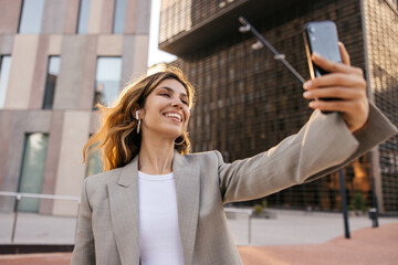 Happy young caucasian girl uses modern phone spending leisure time outdoors. Brown-haired woman wears classic style of clothing. Technology concept