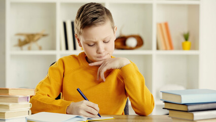 thoughtful preteen schoolboy writing in notebook near books on desk.