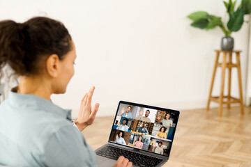 Curly woman waving at the laptop screen with people profiles, using an application for distance video communication with coworkers, webinar participants, meeting online in pandemic