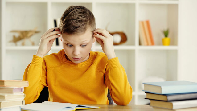 tensed preteen boy looking at notebook while doing homework near books on wooden desk.