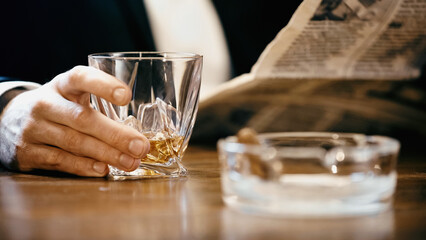 Cropped view of businessman holding glass of whiskey and newspaper near ashtray isolated on black.
