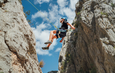 Climber teenager boy in protective helmet abseiling from vertical cliff rock wall using rope Belay...