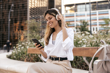 Cheerful young caucasian girl listens to music using headphones and smartphone sitting on bench. Blonde wears classic style of clothing. Audio technology concept