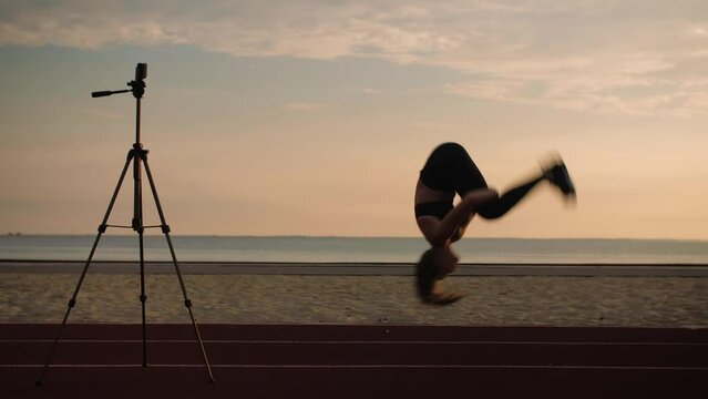 Young Woman in sportwear doing fitness online course.Girl doing back flip, acrobatic activity at sunrise. Woman teaching fitness online on the beach.