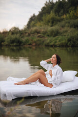 Brunette woman with short hair in a white shirt in bed on the water. A young woman floats on an inflatable mattress with a white sheet on the lake.