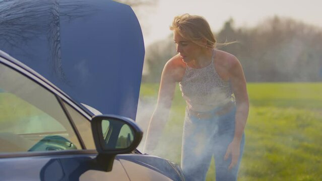 Frustrated woman on country road standing next to broken down car with bonnet up and steam coming out of engine - shot in slow motion