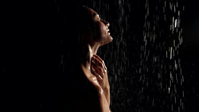 a naked woman stands in the rain and touches neck with hands. dark key. black background. profile view