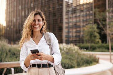 Fototapeta na wymiar Cute young caucasian girl holding smartphone standing outdoors near building. Blonde wears white shirt and backpack. City life concept