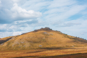 Fototapeta premium Dramatic mountain landscape with sunlit large hill with sharp rocks and trees in cloudy sky. Big shadows of clouds on rocky mountain top. High orange hill in changeable weather in faded autumn color.