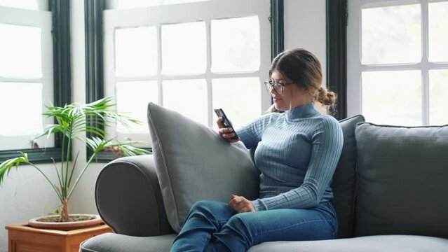 Young beautiful woman sitting on the sofa at home chatting and surfing the net. Female person having fun with smartphone online. Portrait of girl smiling using cellphone revising social media
