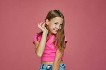 Smiling embarrassed shy caucasian child girl fixing her hair posing on pink studio background....