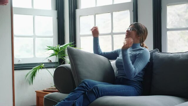 Happy young woman in eyeglasses relaxing on sofa with hands behind head in the living room of her house. Carefree and satisfied lady with torn jeans relaxing on couch at modern apartment