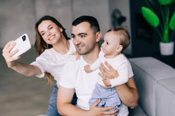 Fototapeta premium Family, parenthood and people concept. Happy mother and father with baby taking selfie at home