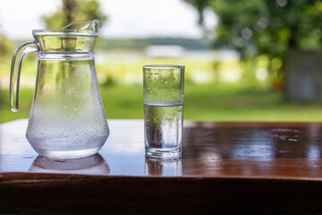 A jug of water and a glass of cold water are placed on a wooden table against a beautiful backdrop.