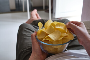 A man eats crispy potato chips from a transparent bowl on the couch. Quick snack. Calories and diet
