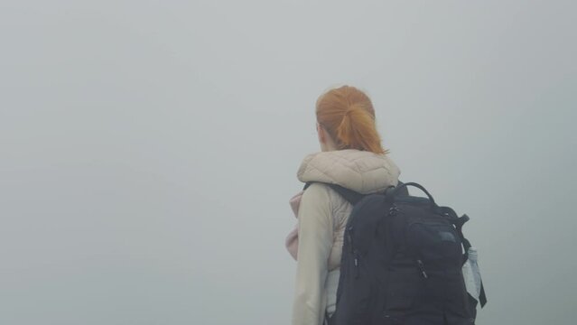 Woman travaler with backpack stops in front of morning mist mountains peak. No landscape view but fog around and female dissapointment. Nature concept.