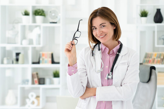 Portrait photo of young and beautiful smiling female doctor in white medical gown standing in the office of the modern clinic