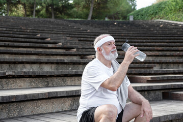 Elderly male retiree sitting on the park stairs drinking water after workout. Concept of active and healthy life in retirement