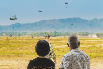 Back view of Asian parents look with worry and concern during parachute training from airplane for army cadet with blurred image of parachute and landscape in background. Family relationship concept.