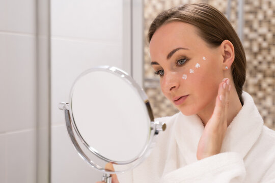 Young woman puts moisturizing cream on face and smears over cheeks looking in mirror. Lady stands in hotel bathroom taking care of skin close view