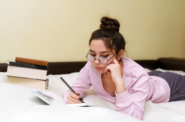 A young woman in glasses lies on the bed and writes information about study or work in a diary, notebook with a pen