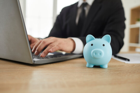 Close up of a blue piggy bank on the office desk of a businessman working on a modern laptop computer in the background. Business, finance, investment, profit concepts