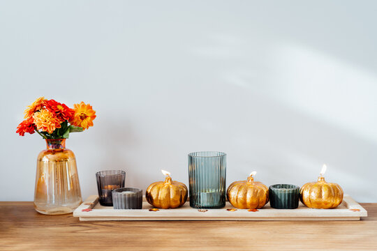 Autumn, fall cozy composition. Fresh dahlia flowers in vase and pumpkin shaped golden candles on the wooden tray with white wall background. Scandinavian minimalist hygge home decor. Selective focus