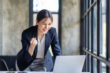 Happy excited successful Asian businesswoman triumphing with a laptop computer smartphone in the workplace office