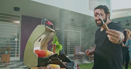 Barbecue chef prepares meat during summer garden party. Portrait of man slicing sausage showing to camera. Happy person preparing food