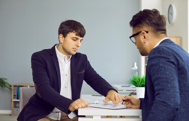 Two serious business people in office sign a contract sitting at desk, manager gives document to...