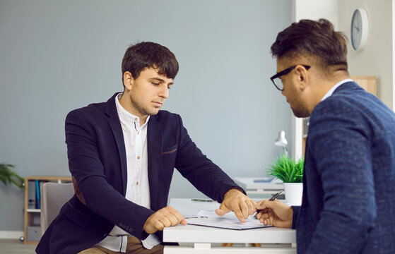 Two serious business people in office sign a contract sitting at desk, manager gives document to his client. Business meeting, transaction, conclusion of contract, provision of services concept.