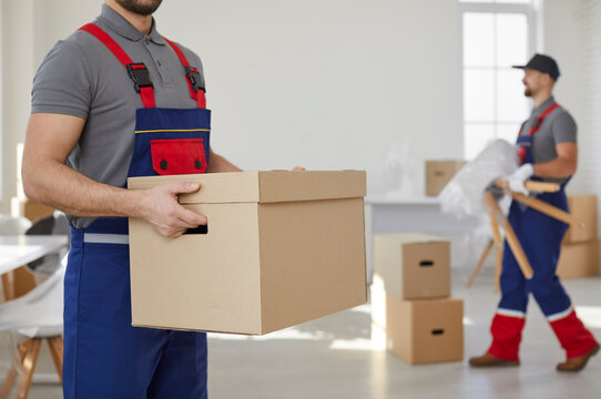 Moving service. Workers of transport and delivery company transport cardboard boxes and furniture. Close up of cardboard box in hands of man in coveralls on background of his colleagues. Cropped image