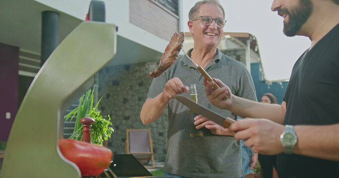 Father Standing By Son Grilling Meat. Adult Son Showing Steak To Dad. Friends And Family Gathered For Barbecue. Parent Talking To Son During Summer Holiday Celebration