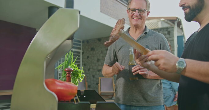 Father Standing By Son Grilling Meat. Adult Son Showing Steak To Dad. Friends And Family Gathered For Barbecue. Parent Talking To Son During Summer Holiday Celebration