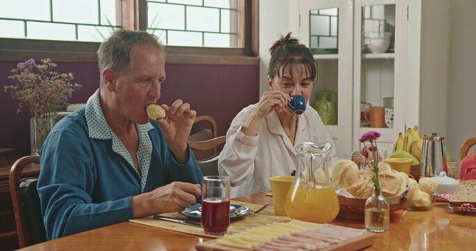 A Middle Aged Couple Chatting During Breakfast. They Are Sitting At The Lavish Kitchen Table, Smiling And Wearing Pajamas. Married Couple In Conversing In The Morning Breakfast Table