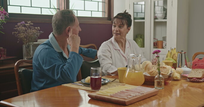 A Middle Aged Couple Chatting During Breakfast. They Are Sitting At The Lavish Kitchen Table, Smiling And Wearing Pajamas. Married Couple In Conversing In The Morning Breakfast Table