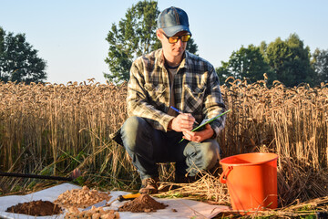 Male agronomist preparing soil samples for laboratory analysis, writing in information sheet outdoors at sunrise. Man farmer taking notes in form, working at field. Soil certification