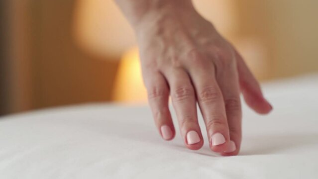 Hand of woman with neat manicure touches white clean sheet put on bed on blurred background. Guest enjoys facilities in hotel room closeup slow motion