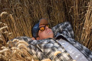 Young adult farmer relaxing at agricultural grain field at sunny day. Male worker wearing hat, checkered shirt, sunglasses, lying in ripe wheat field outdoors. Countryside concept. Simple living - obrazy, fototapety, plakaty