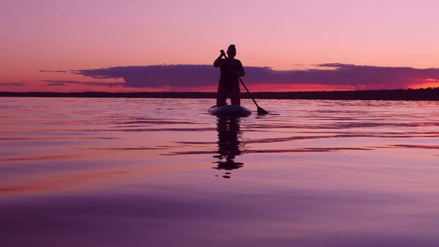 A woman in a swimsuit silhouette floats on a SUP board on the water at sunset twilight against a purple sky reflection in the water. Overall plan