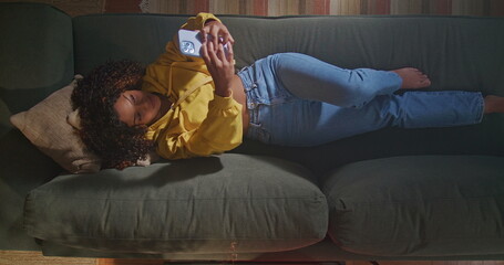 African American woman lying on sofa holding smartphone seen from above angle. A black girl taking selfie with phone