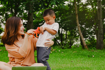 Mother beautiful sits in the garden with her asian son playing T-rex dinosaur dolls, playful toy...