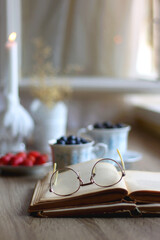 Vintage tea set filled with blueberries, blackberries and starwberries, open book, reading glasses and decorative candles on the table. Selective focus.
