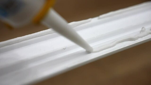 Close up shot of applying white glue on plastic surface of decorative baseboard, craftsman using tube with adhesive glue for fixing plastic plinth to the wall. Skirting boards gluing process, final