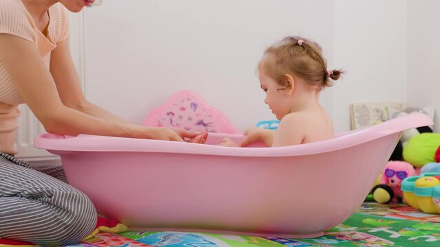 Side view mother daughter baby in bathroom mothers mouth is talking to child, they are pouring water from their hand into cup. Kid listen carefully to mother repeats the actions, mother is happy.