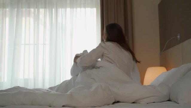Woman wakes up in early morning stretching arms in hotel room. Female guest gets out of bed coming to bright window covered with blanket backside view