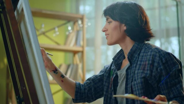 A beautiful woman with a funky pixie haircut is painting something on a large canvas and wearing a blue flannel top that has paint marks on it while being in an art studio