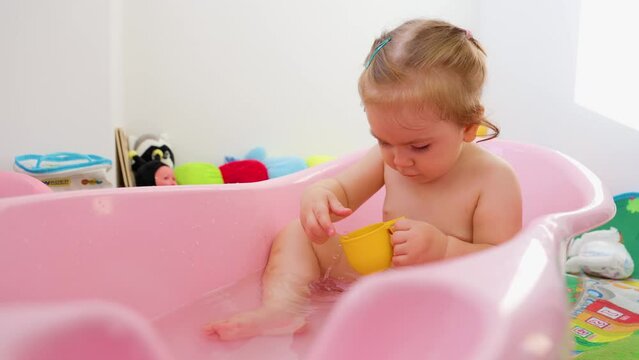 Front view playing happy little girl bathing in a baby pink bath attentively playing with a small toy and a mug. The child watches with interest how the toy thinks in the water and takes it out.