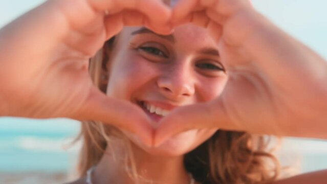 Young beautiful Caucasian woman looking at camera through heart made of hands with smile saying hello to friends from vacation or exotic resort stands on beach next to ocean. Lifestyle, positive