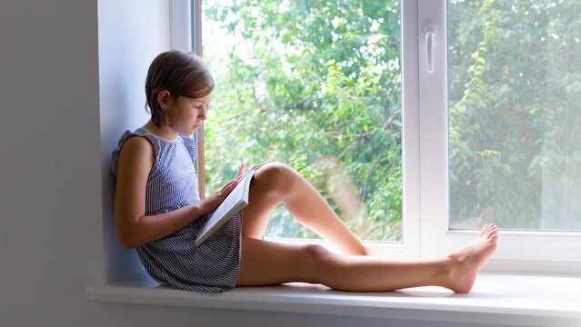 Side view of pretty child serious read book sitting on windowsill moving toes of one leg other bent at knee. Turns the pages of the book with interest. Background light of walls, windows, sunlight.